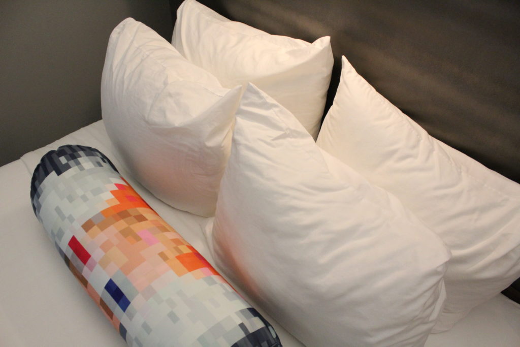 4 white pillows on a hotel room bed with a vanity pillow with pixelated cartoon character at the Aloft Las Colinas hotel