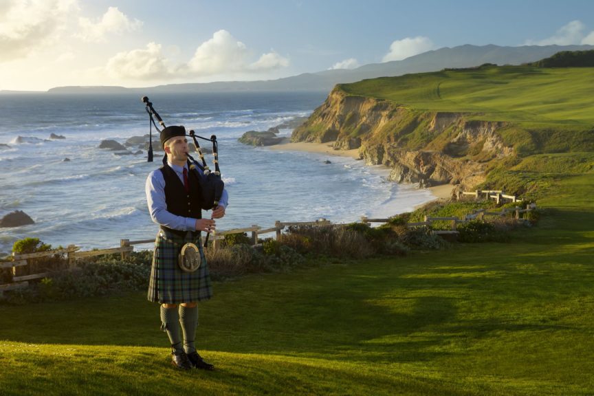 a man playing bagpipes on a grassy hill overlooking a body of water