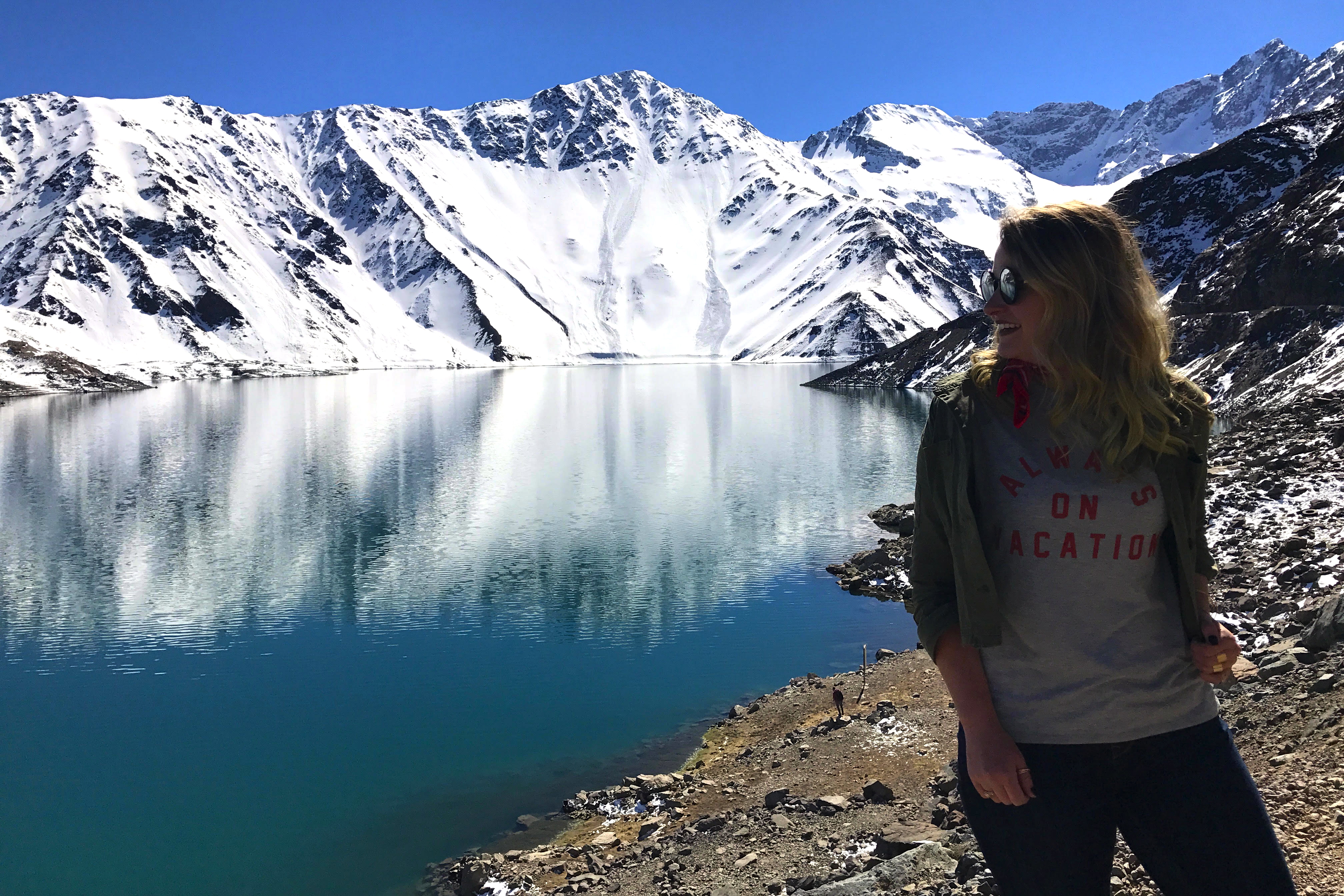 a woman standing next to a body of water with snow covered mountains in the background