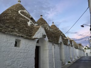 a row of white houses with a few pointed roofs with Alberobello in the background