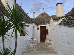 a white stone building with a door and a table with Alberobello in the background