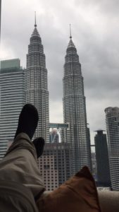 a person's foot in front of a tall building with Petronas Towers in the background