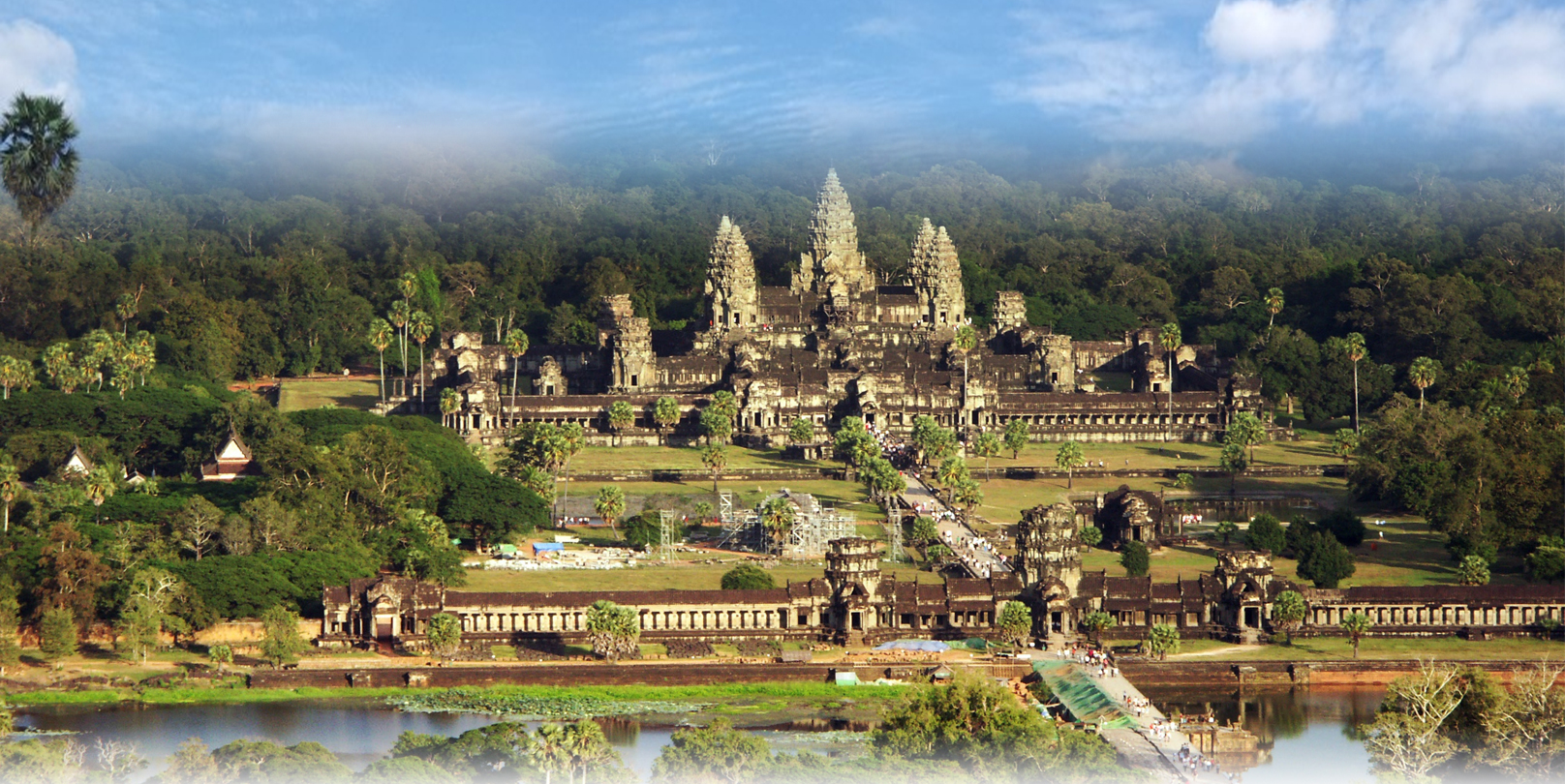 a large stone building with trees and a river with Angkor Wat in the background