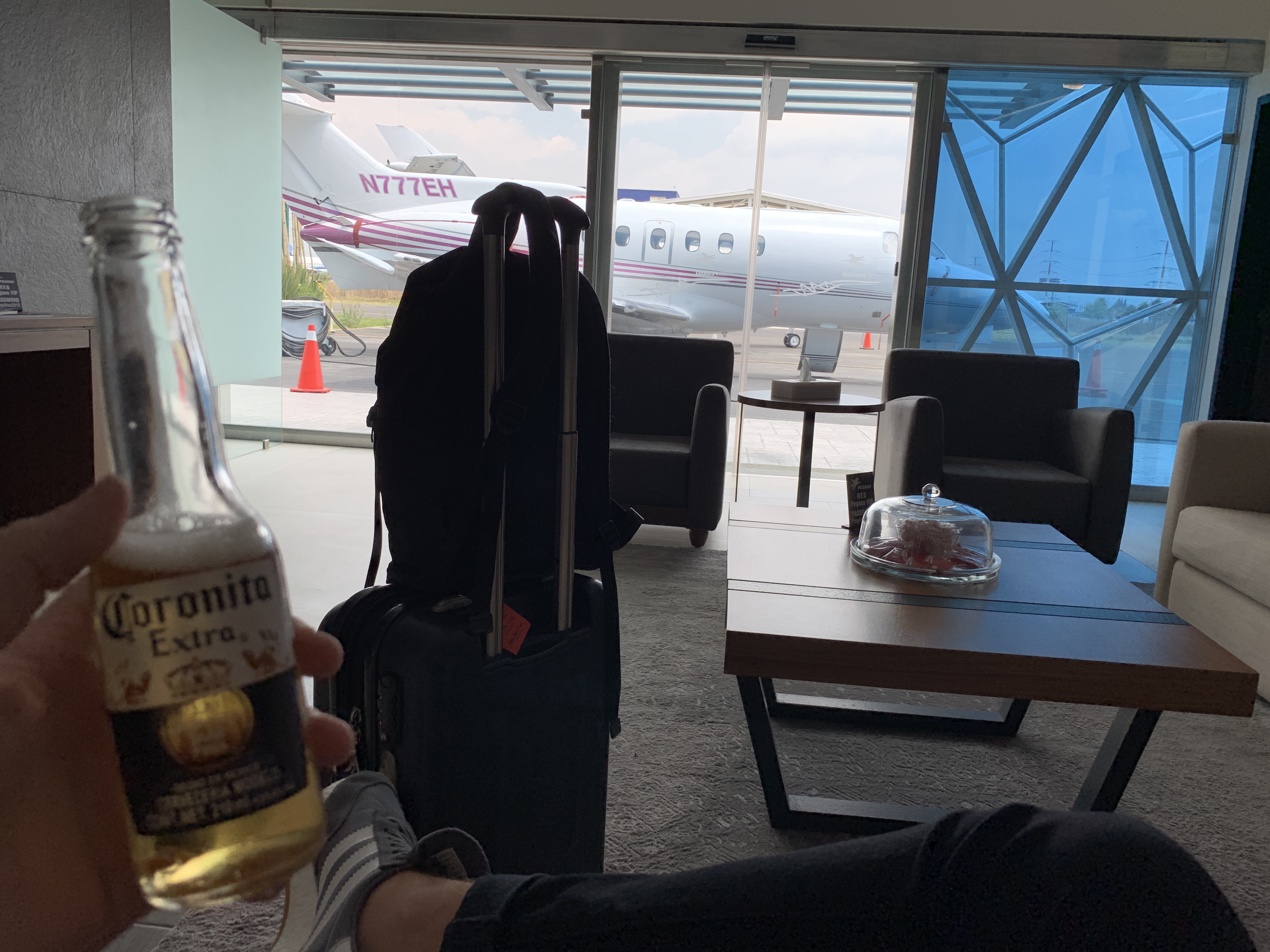 a hand holding a bottle of beer and luggage in front of an airplane
