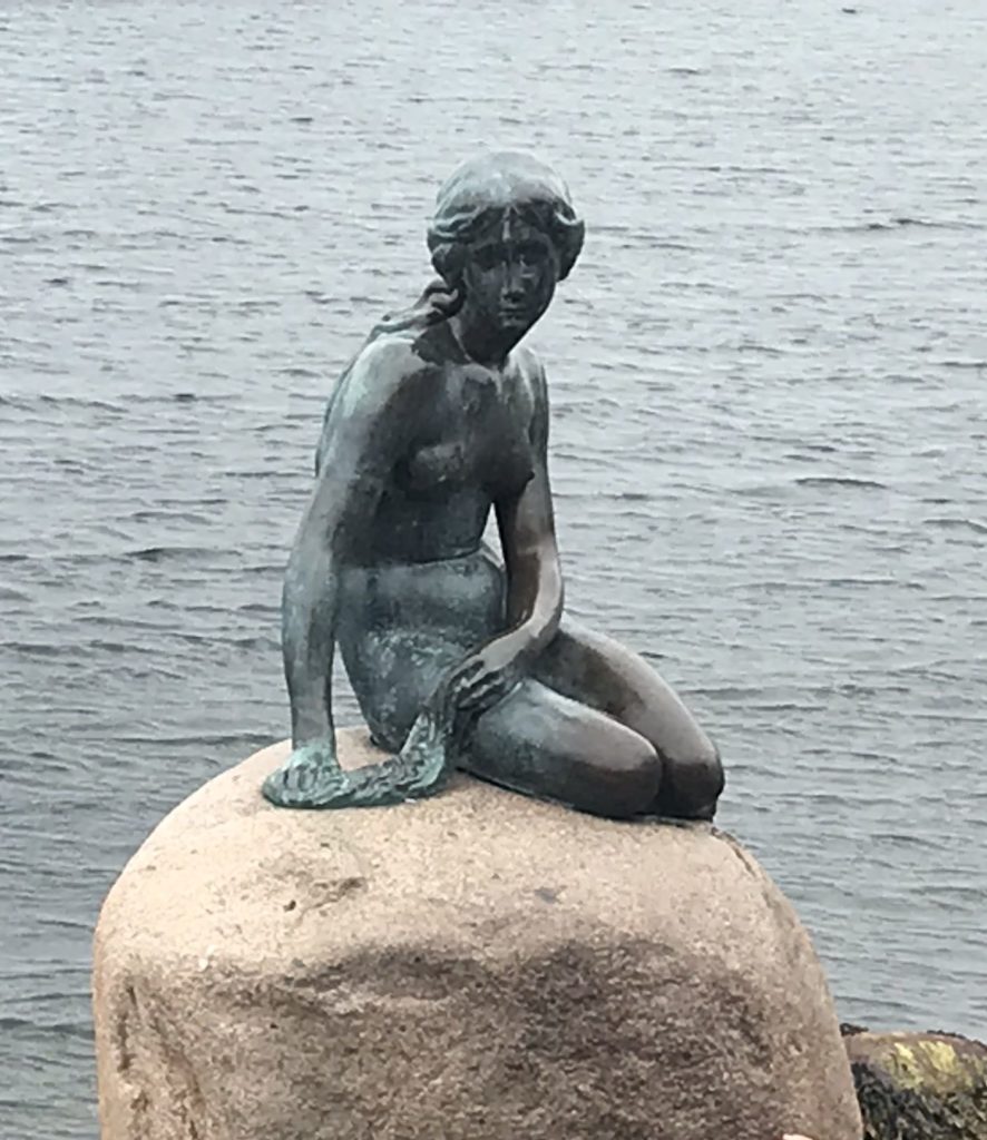 a statue of a woman sitting on a rock with The Little Mermaid in the background