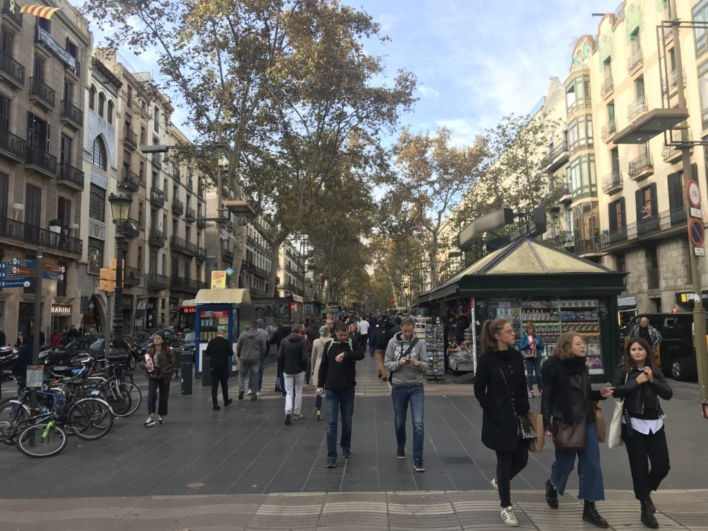 a group of people walking on a sidewalk with La Rambla, Barcelona in the background