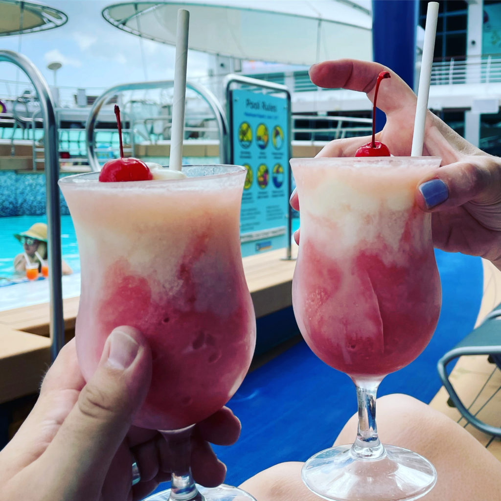 two hands holding drinks with straws and a pool in the background
