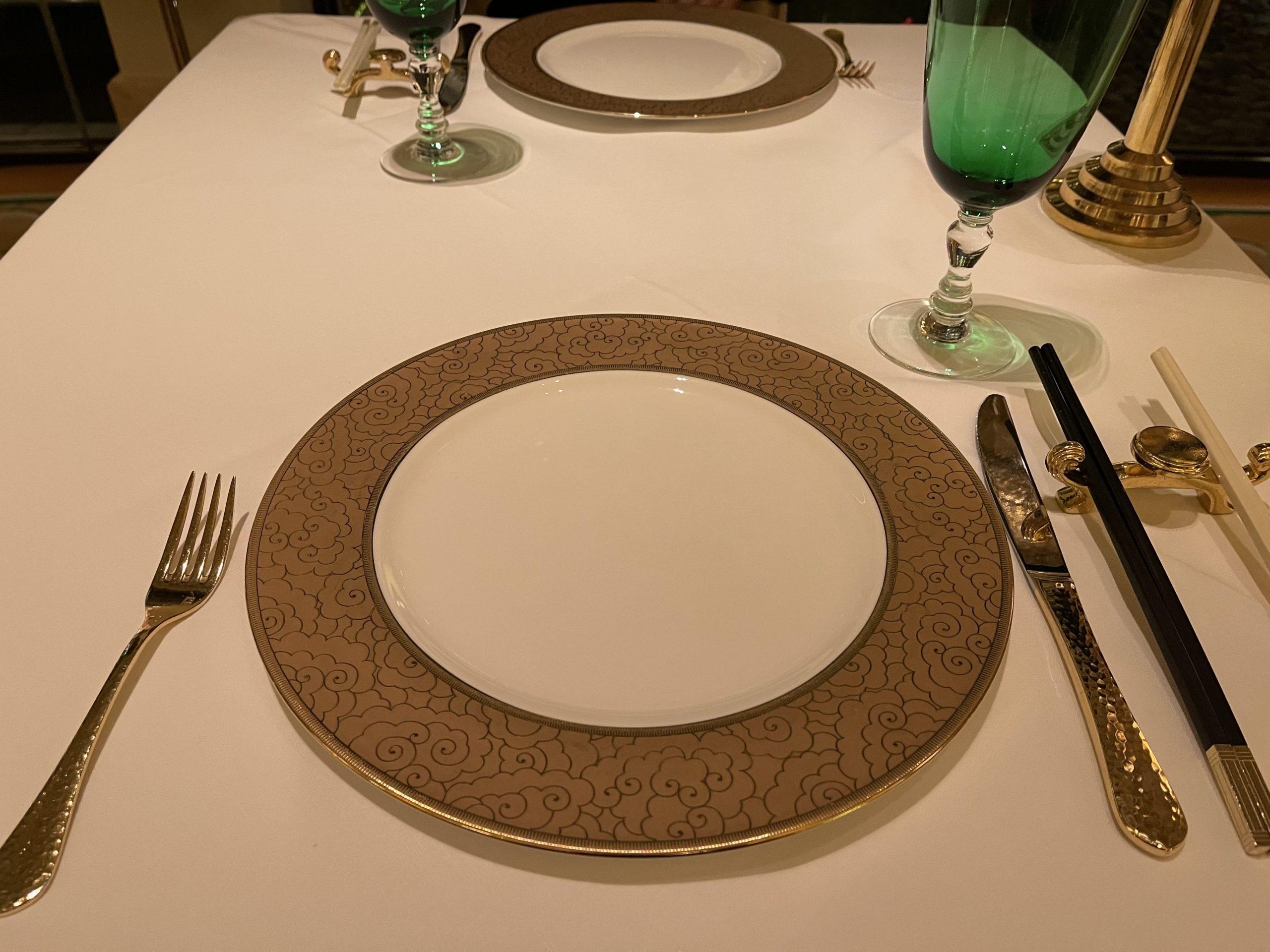 a plate with a gold rim and a gold rim on a table with a green glass and a silverware