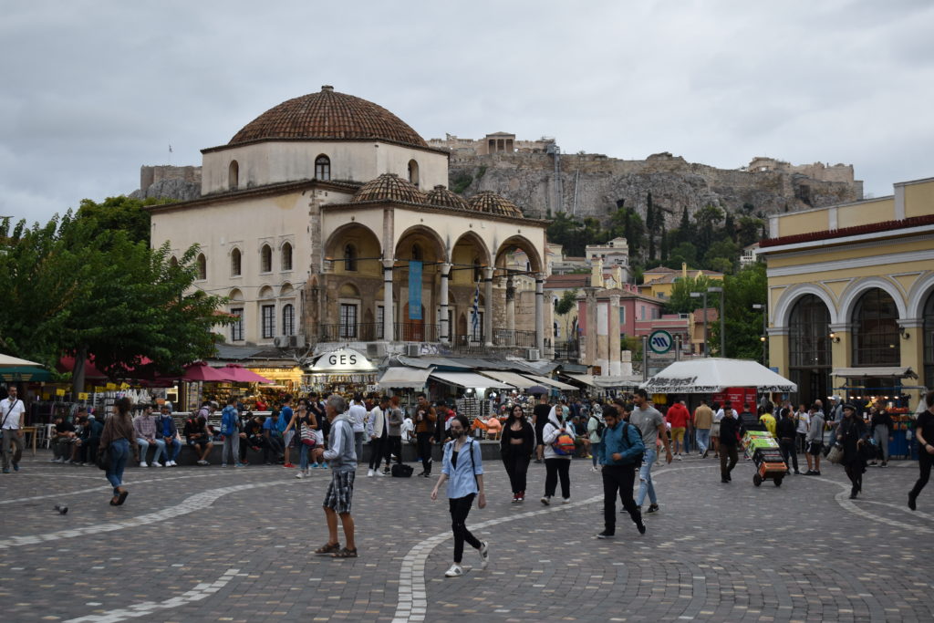 a group of people in a square with a dome shaped building in the background with Monastiraki in the background