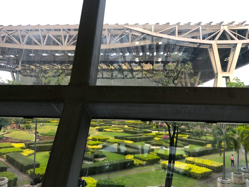 View of the outside garden from the window in the Coral Lounge. The gardens have paths between the greenery. A person is seen walking.