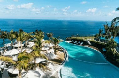 A hotel pool with palm trees and a beach next to a blue body of water