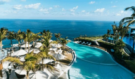 A hotel pool with palm trees and a beach next to a blue body of water