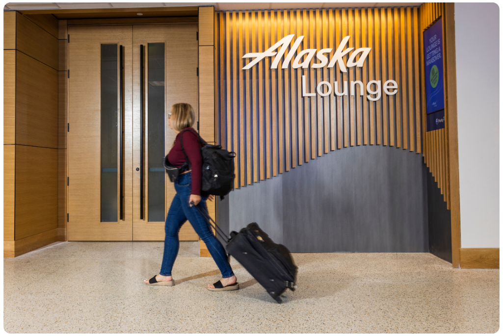 A woman walking with luggage in front of an Alaska Lounge airport club