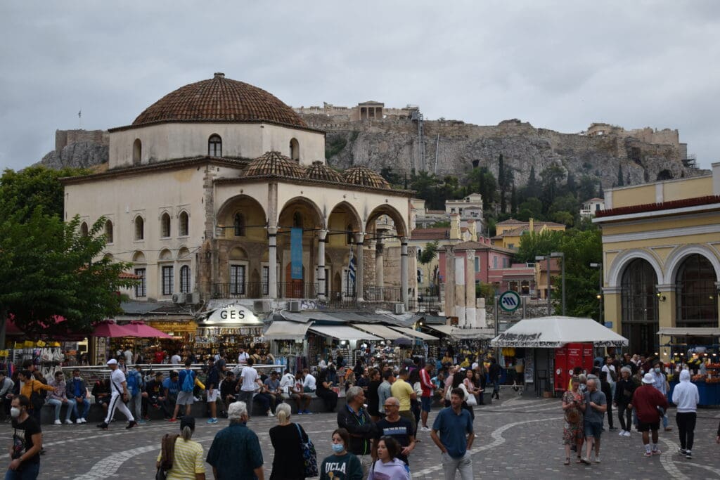 a group of people in a plaza with a dome shaped building with Monastiraki in the background