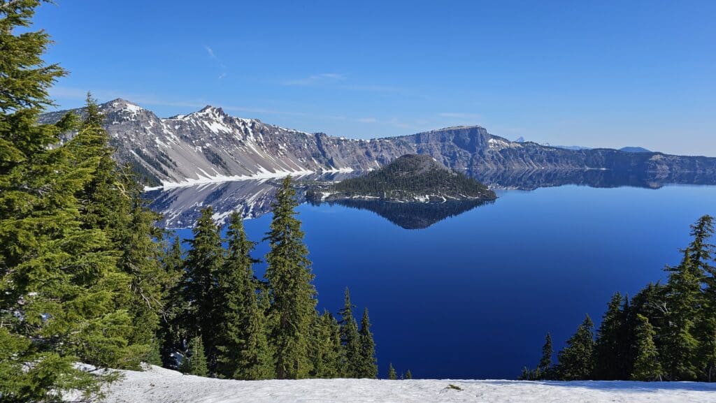 a snow covered mountain with trees and a body of water with Crater Lake National Park in the background