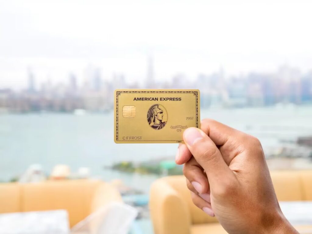 A person hold up an Amex Gold Card against the backdrop of a city skyline