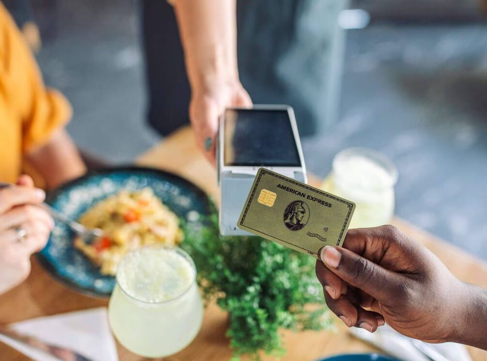 A person at a restaurant table holding an American Express Gold card over a card reader