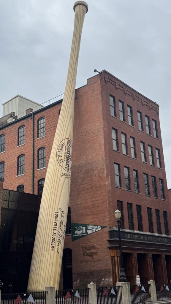 a large wooden bat in front of a brick building with Louisville Slugger Museum & Factory in the background